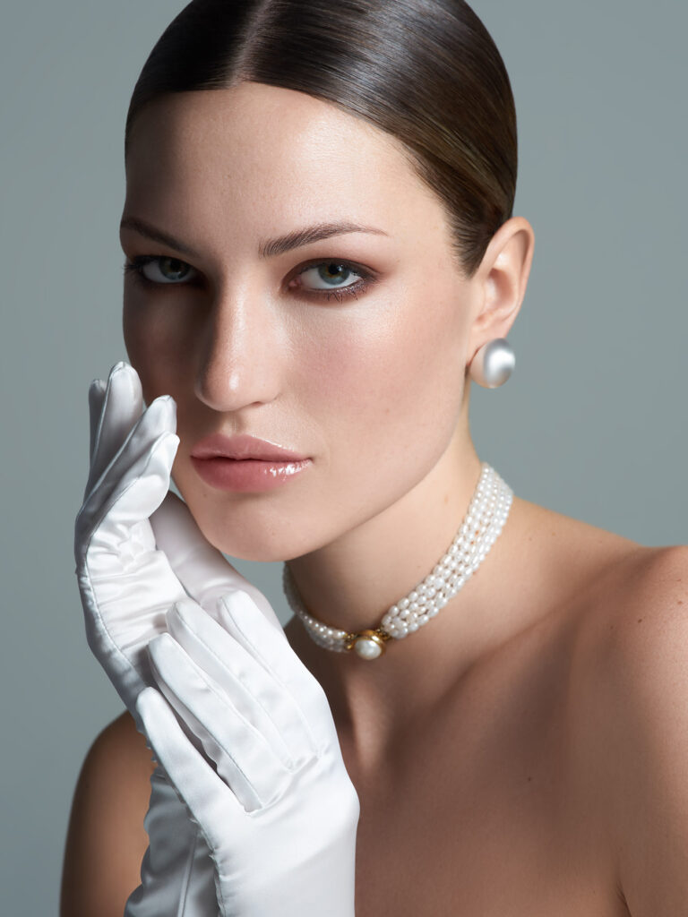 Portrait of a woman in bridal attire with sleek brown hair pulled back, wearing white gloves, pearl stud earrings, and a multi-strand pearl necklace, with one gloved hand positioned near her face against a neutral background