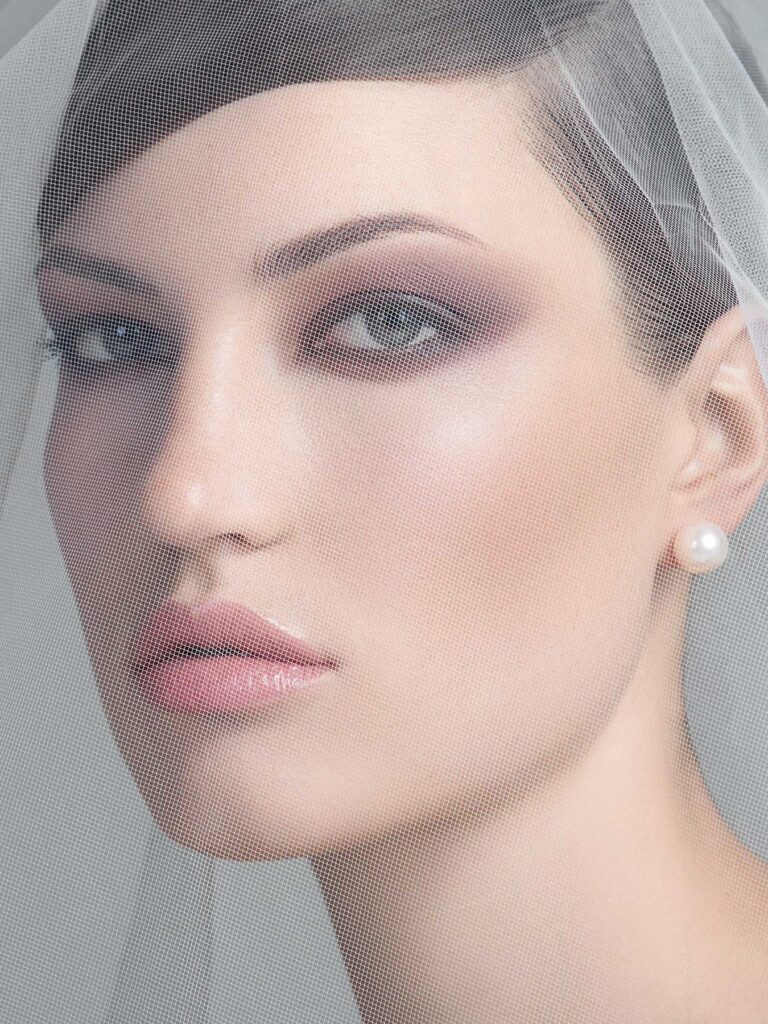 Close-up portrait of a bride wearing a white wedding veil that partially covers her face, with natural makeup, pearl earrings, and a gentle smile looking upward