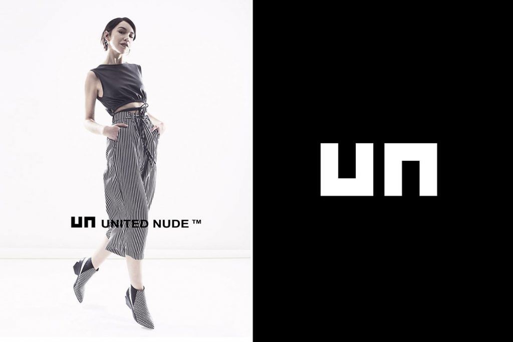 Fashion photography of a model dressed in a simple outfit of charcoal sleeveless top wrapped around the waist and striped high-waisted wide-leg pants with perforated ankle boots. The model is posing in front of a divided background - white to the left, black to the right - with the 'UN UNITED NUDE™' logo centered over both segments