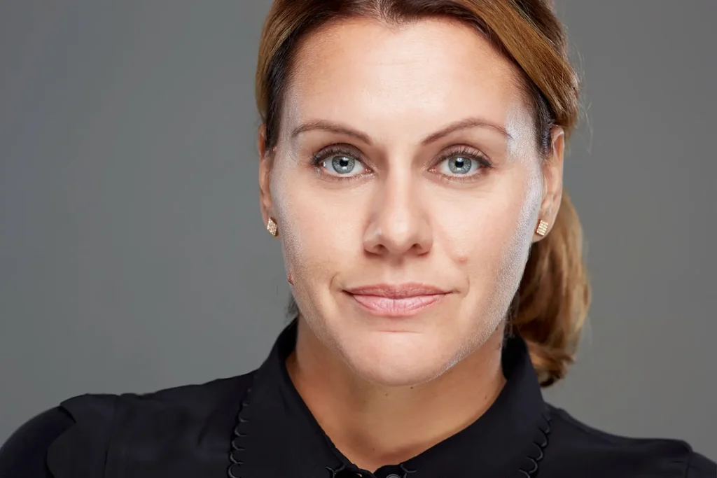 Professionelles Portrait einer Frau mit zurückgekämmten blonden Haaren und blauen Augen, trägt goldene Ohrringe und ein schwarzes Hemd, aufgenommen vor neutralem grauem Hintergrund.