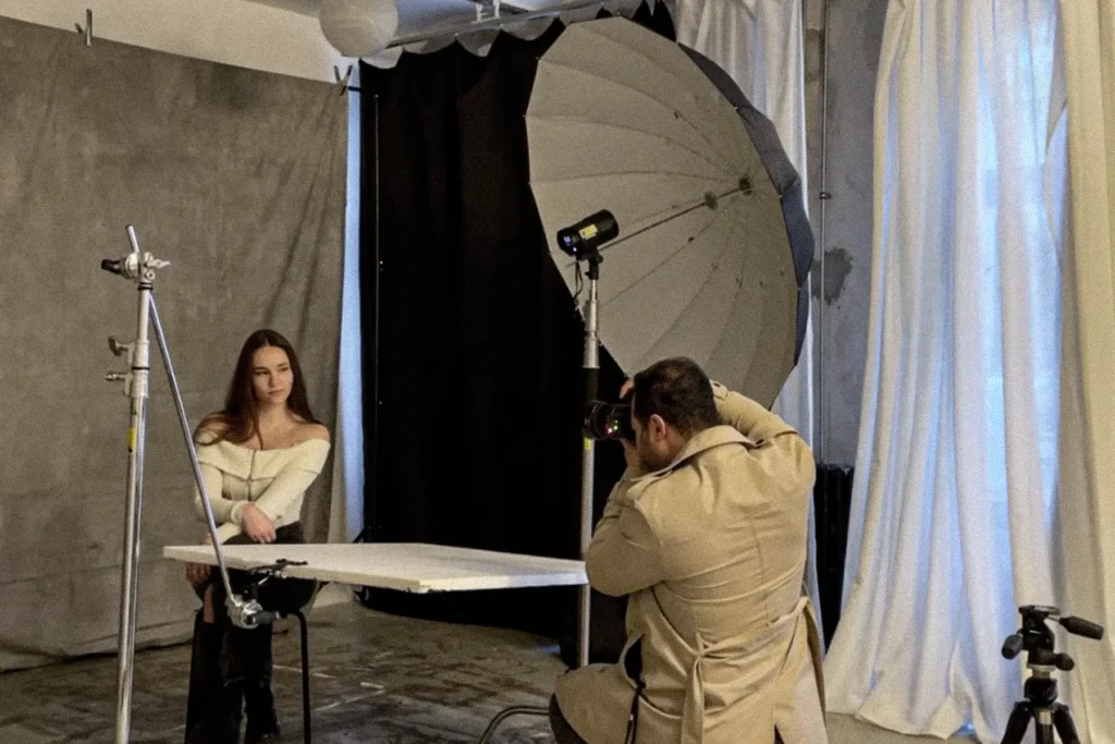 Blick hinter die Kulissen einer professionellen Sedcard-Fotosession in einem Berliner Studio, zeigt einen Fotografen in beigem Hoodie beim Fotografieren einer weiblichen Model in schulterfreiem weißem Oberteil, mit professioneller Beleuchtungsausrüstung einschließlich Schirmen, Reflektoren und Studioblitzen vor drapierten Hintergründen