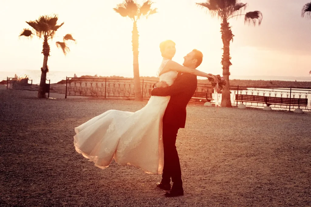 A bride and groom embrace in a romantic dance pose on a beach promenade at sunset.