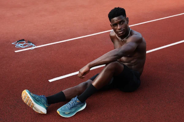 A young Black male athlete sits on a red running track in Berlin Ostkreuz (VSG Weberwiese 72), wearing black athletic shorts and teal/blue HOKA running shoes. He is looking directly at my camera.