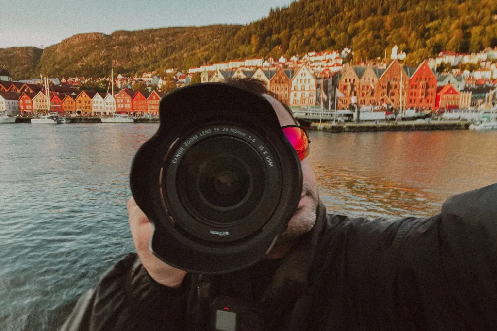 Photographer Ed Mehravaran, wearing a red cap and dark jacket, holds a large Canon camera lens to his eye and photographs Bergen's famous Bryggen harbor district. The scene shows the iconic row of colorful wooden houses along the harbor—traditional Hanseatic architecture in bright shades of red, orange, yellow, and white, reflected in the calm water.