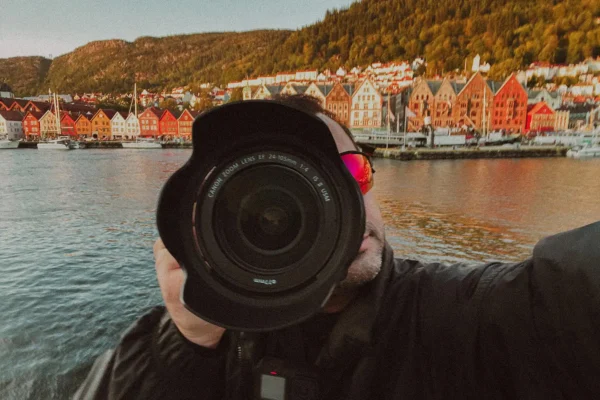 Photographer Ed Mehravaran, wearing a red cap and dark jacket, holds a large Canon camera lens to his eye and photographs Bergen's famous Bryggen harbor district. The scene shows the iconic row of colorful wooden houses along the harbor—traditional Hanseatic architecture in bright shades of red, orange, yellow, and white, reflected in the calm water.
