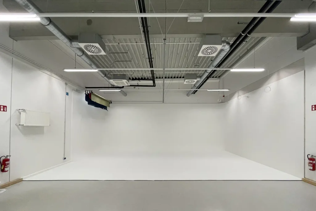A large, empty white photography studio or cyclorama with seamless white walls and floor. The exposed industrial ceiling features fluorescent lighting strips, ventilation grilles, black ductwork, and mechanical equipment. A professional photography backdrop system is mounted on the left wall, and red fire extinguishers are visible in both corners. The space has a clean, minimal aesthetic typical of commercial photo studios.