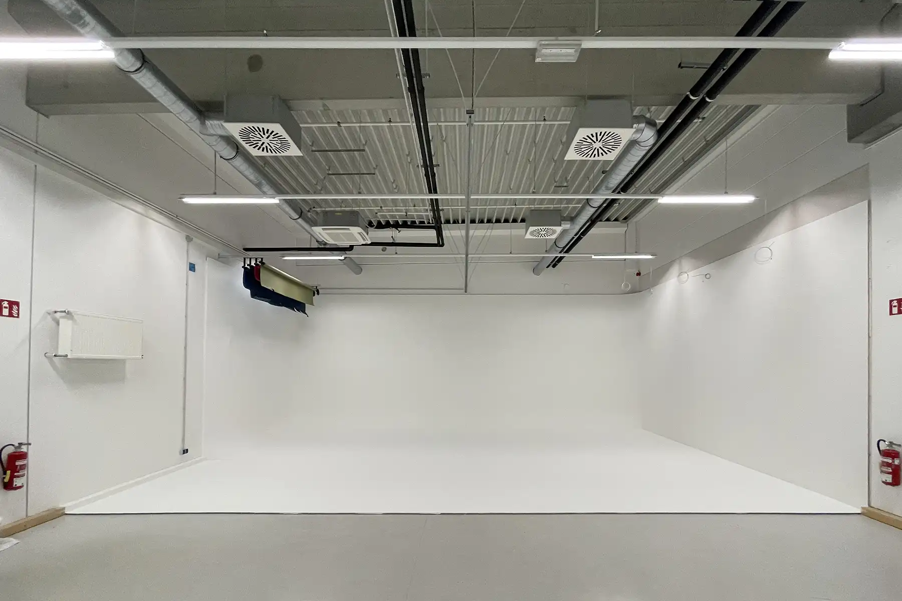 "A large, empty white photography studio or cyclorama with seamless white walls and floor. The exposed industrial ceiling features fluorescent lighting strips, ventilation grilles, black ductwork, and mechanical equipment. A professional photography backdrop system is mounted on the left wall, and red fire extinguishers are visible in both corners. The space has a clean, minimal aesthetic typical of commercial photo studios."