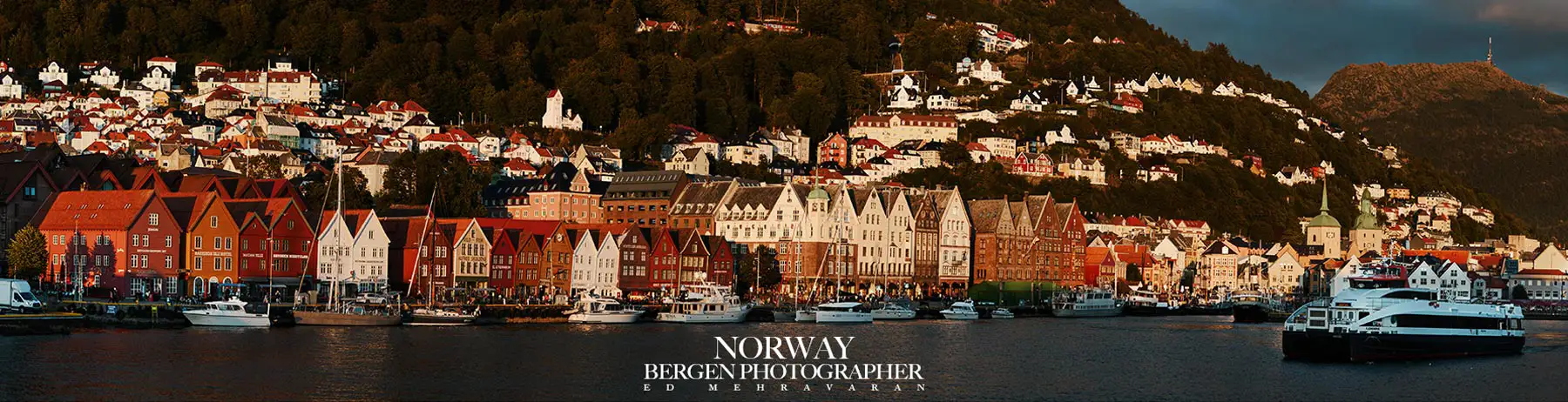 This panoramic photo shows the historic Bryggen harbor district in Bergen, Norway. It shows the striking row of colorful wooden houses along the waterfront promenade. They are characterized by traditional Hanseatic architecture with their distinctive pointed gables and bright facades in shades of red, orange, yellow, and white. The buildings are reflected in the calm harbor waters, where several boats and yachts are moored. Behind the historic buildings, residential houses nestle up the hills, nestled in lush green forests. The scene is bathed in warm, golden light that highlights the rich colors of the buildings and offers a picturesque view of this UNESCO World Heritage Site. At the bottom of the image is the text “NORWAY BERGEN PHOTOGRAPHER Ed Mehravaran.”