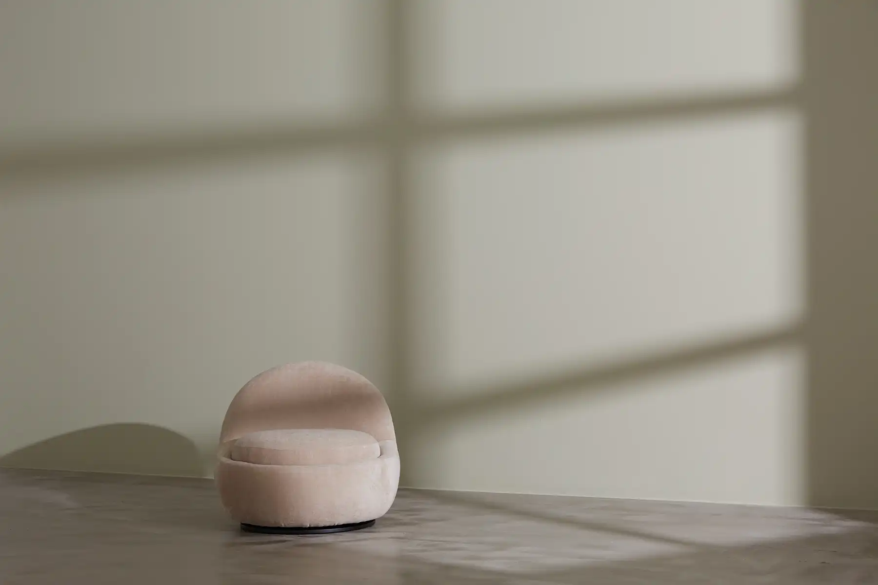 A soft, rounded pink velvet ottoman or pouf sits on a polished concrete floor in a minimalist interior space. The background shows blurred white walls with geometric window shadows creating subtle rectangular patterns. The scene has a dreamy, soft-focus quality with warm, diffused lighting.