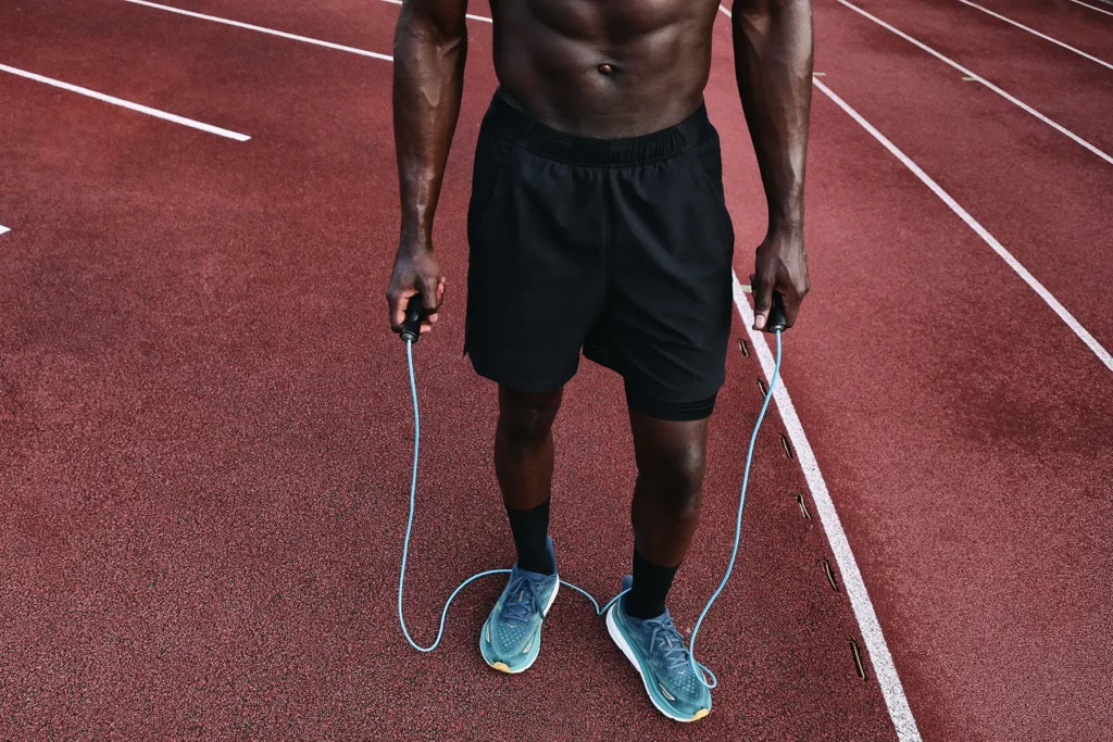 Athletische Mann mit definierten Bauchmuskeln auf einer roten Laufbahn haltend und umklammernd ein blaues Springseil in den beiden Händen, schwarze Shorts tragend und blaue Laufschuhe an den Füßen, mit weißen Spurlinien der Bahn im Hintergrund