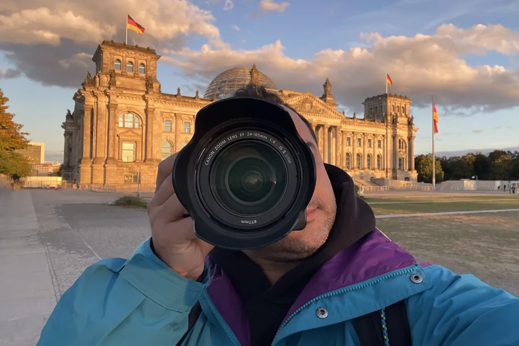 Ein Fotograf fotografiert mit einer Canon Kamera und einem Zoomobjektiv (EF 24-105mm) vor dem Reichstagsgebäude in Berlin. Der Fotograf trägt eine türkisfarbene Jacke und einen lila Kapuzenpullover. Hinter ihm ist das historische Reichstagsgebäude mit seiner charakteristischen Glaskuppel und wehenden Deutschlandflaggen im goldenen Abendlicht unter einem leicht bewölkten Himmel zu sehen.