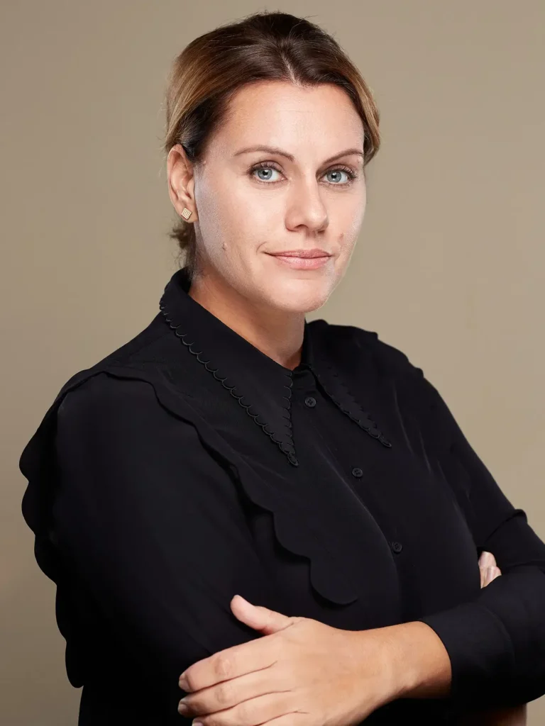 Businessportrait einer Frau in Berlin mit geschlossenen blonden Haaren und schwarzem Hemd, die die Arme verschränkt und in die Kamera blickt. Der Hintergrund ist beige.