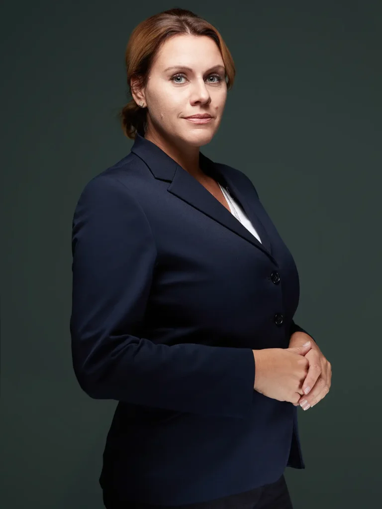 Businessporträt einer Frau in Berlin mit geschlossenen blonden Haaren, einem marineblauen Blazer und weißem Hemd, die ihre Hände hält und in die Kamera blickt. Der Hintergrund ist dunkelgrün.