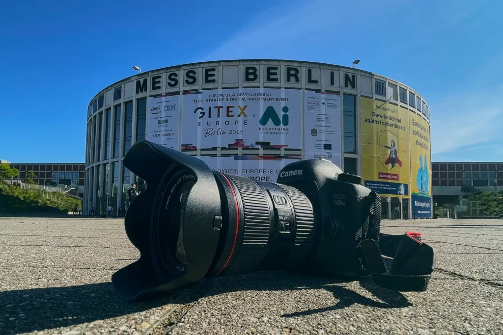 Canon-Kamera eines Eventfotografen mit aufgesetztem Objektiv auf dem Boden vor dem Messezentrum Berlin, mit einem großen Banner für die GITEX Europe Berlin 2025, Europas größte Messe für Technologie und Start-ups.