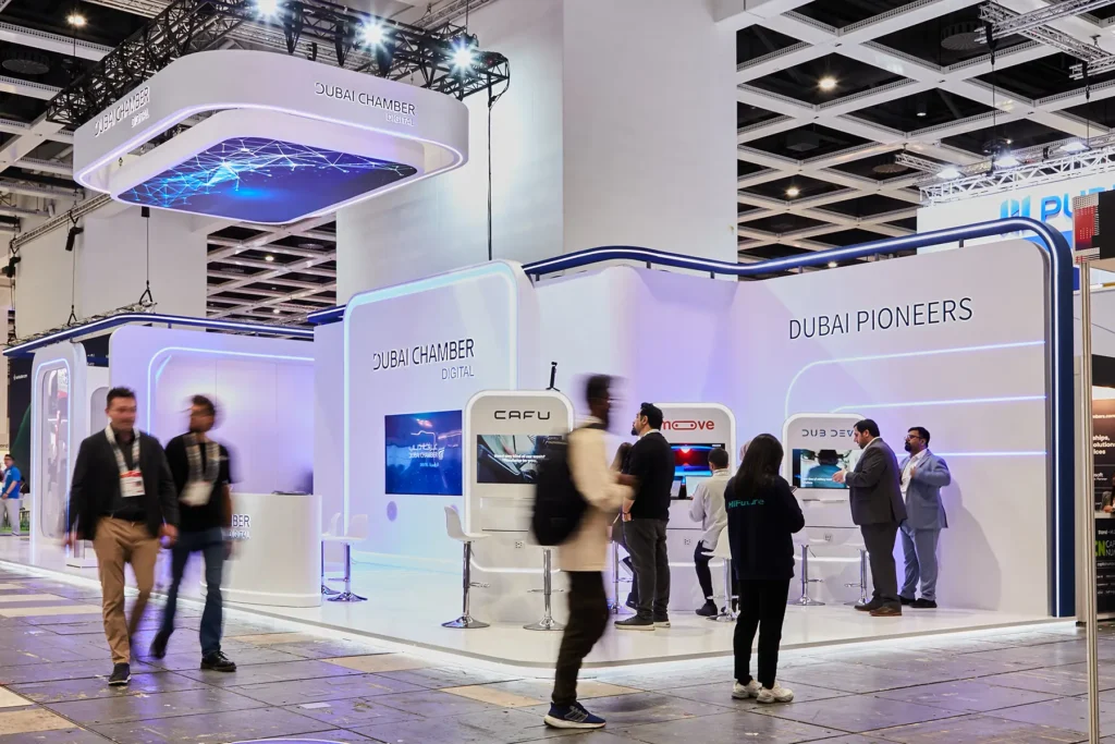 Fotografie auf der Messe Berlin für einen Technologie-Messestand für Dubai Chamber Digital und Dubai Pioneers auf einer Fachmesse, mit weißer, geschwungener Architektur mit blauer LED-Akzentbeleuchtung, über Kopf hängenden Bildschirmen mit Netzwerkgrafiken und Menschen, die vorbeigehen und sich im Standbereich vernetzen.