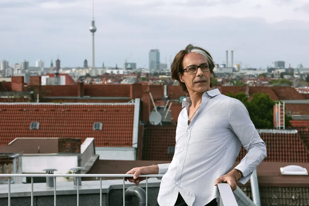 Porträt von Arian. Er ist ein Mann mittleren Alters mit braunen Haaren und Brille. Auf diesem Bild trägt er ein hellblaues Hemd. Im Hintergrund sieht man die Berliner Skyline, und der Fernsehturm ist im Bild sichtbar.