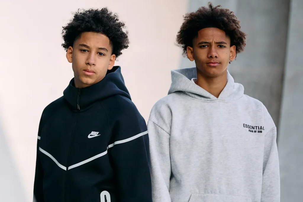 Auf diesem Sedcard-Foto sieht man zwei junge Teenager mit lockigen Haaren, die nebeneinander vor einem neutralen Hintergrund stehen. Es sind Zwillinge aus Anny. Der Junge links, Xavier, trägt einen schwarzen Nike-Hoodie mit weißen Chevron-Streifen auf der Brust, während der Junge rechts, Levi, einen hellgrauen Hoodie mit dem Aufdruck „ESSENTIALS FEAR OF GOD“ trägt. Beide blicken ernst in die Kamera.