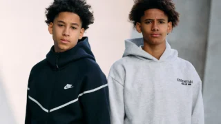 Auf diesem Sedcard-Foto sieht man zwei junge Teenager mit lockigen Haaren, die nebeneinander vor einem neutralen Hintergrund stehen. Es sind Zwillinge aus Anny. Der Junge links, Xavier, trägt einen schwarzen Nike-Hoodie mit weißen Chevron-Streifen auf der Brust, während der Junge rechts, Levi, einen hellgrauen Hoodie mit dem Aufdruck „ESSENTIALS FEAR OF GOD“ trägt. Beide blicken ernst in die Kamera.