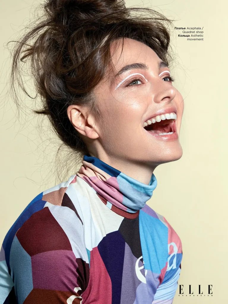 Beauty Portrait of a smiling woman in profile wearing a colorful geometric patterned turtleneck against a pale yellow background, with voluminous brown hair in an updo and artistic white graphic eye makeup, featured in Elle Kazakhstan magazine. This photo was taken by Ed Mehravaran