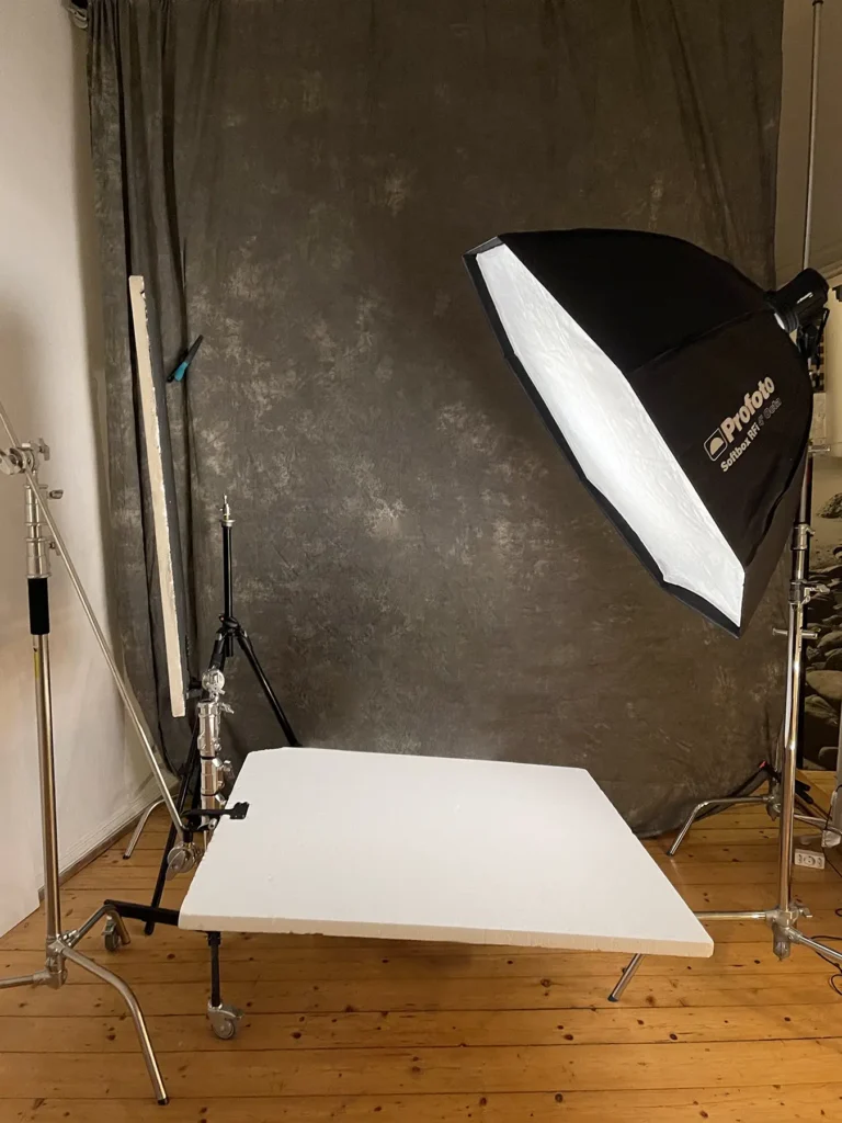 Photography studio setup in Berlin with a white reflector held by a C-stand, illuminated by a Profoto softbox on a stand against a gray textured backdrop, with additional lighting equipment on tripods. This is Ed Mehravaran's photo Studio in Berlin.