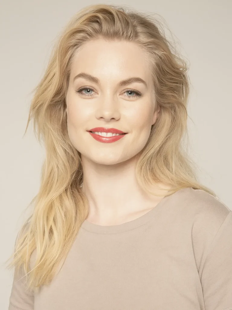Studio portrait of a smiling woman with wavy blonde hair wearing a beige t-shirt and coral-red lipstick against a neutral beige background