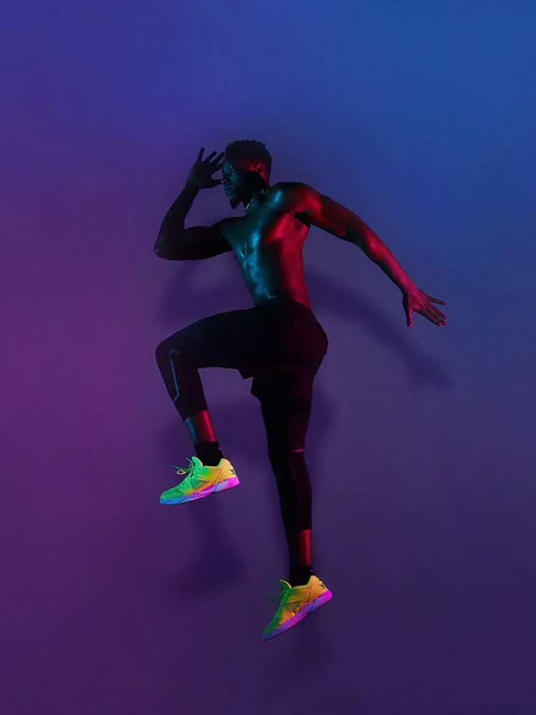 Sport photo of an athletic man jumping mid-air against a purple and blue gradient background, wearing black pants and vibrant rainbow-colored sneakers, with dramatic red and blue lighting illuminating his torso. Ed Mehravaran taked this photos with special lighting teqnic with using color gels. blue color gel in front of flash light from the right side and magenta color gel from the left side of the picture.