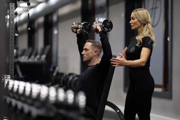 A female personal trainer in Holmes Place Berlin wearing a black outfits. This personal trainer showin to a young man how to do overhead dumbbell shoulder pressses