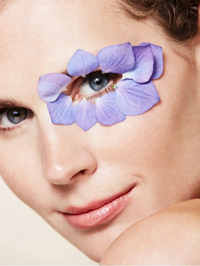 Close-up beauty portrait by Berlin photographer Ed Mehravaran showing a woman with blue eyes framed by purple hydrangea petals, showcasing high-end beauty photography in Berlin by the Professional photographer Ed Mehravaran