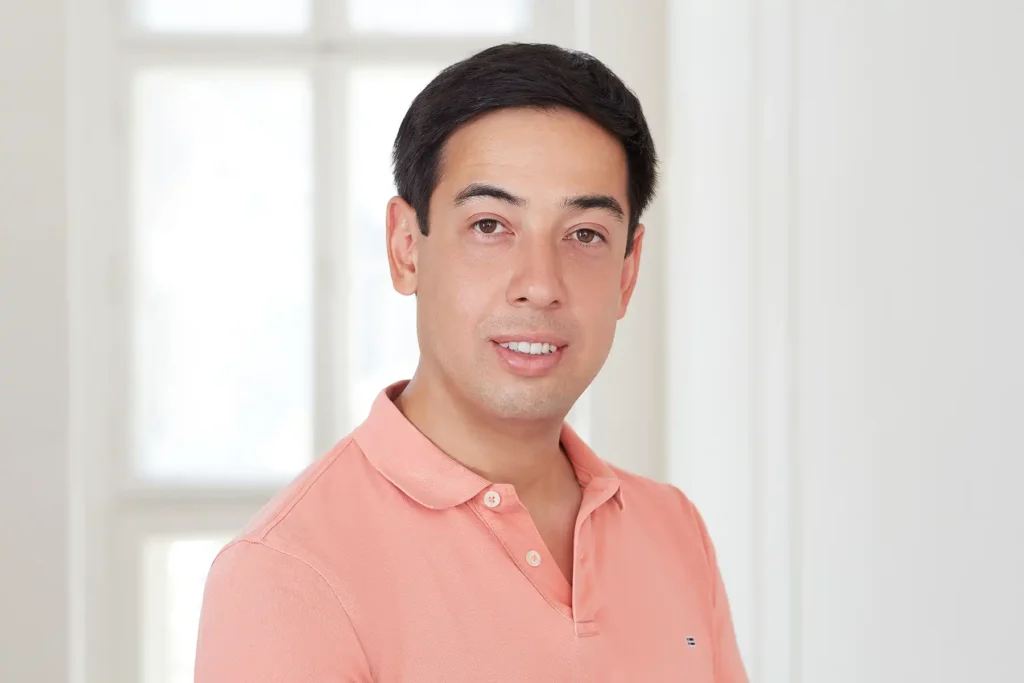 Professionelles Bewerbungsfotos Berlin eines jungen Mannes mit kurzen dunklen Haaren, der ein lachsrosa Poloshirt trägt, vor einem hellen, leicht verschwommenen weißen Studiohintergrund