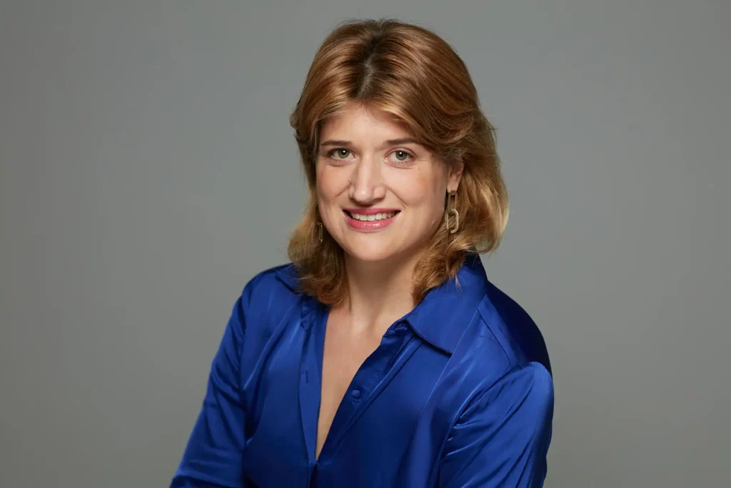 Professionelles Bewerbungsfoto von Rike mit schulterlangem, kastanienbraunem Haar und einem freundlichen Lächeln. Sie trägt eine kobaltblaue Satinbluse und goldene Ohrhänger vor einem neutralgrauen Studiohintergrund.