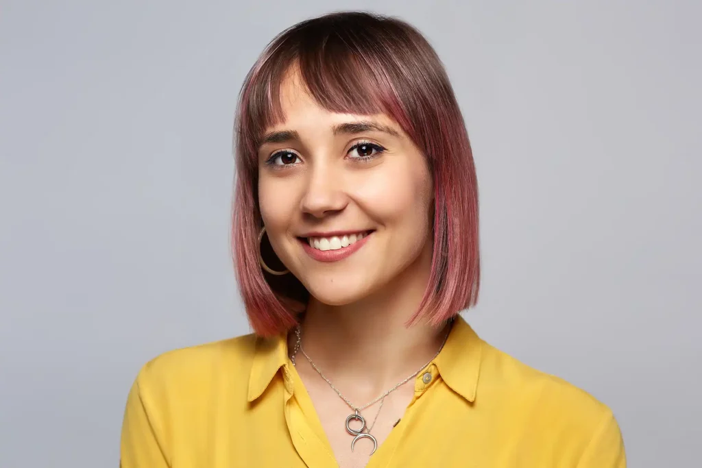 Professionelles Bewerbungsfoto einer jungen Frau mit rosafarbenem Bob und Pony, die ein senfgelbes Hemd und mehrere silberne Halsketten trägt und vor einem neutralgrauen Studiohintergrund selbstbewusst lächelt.