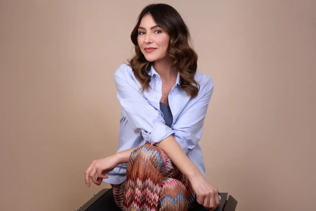 Frau sitzt entspannt auf einem dunklen Hocker vor einem beigen Studiohintergrund. Sie trägt ein hellblaues Leinenhemd mit aufgekrempelten Ärmeln und eine gemusterte Hose mit buntem Zickzackmuster in Rot-, Orange- und Blautönen. Ihre dunkelbraunen, welligen Haare fallen über die Schultern. Sie lächelt leicht in die Kamera.