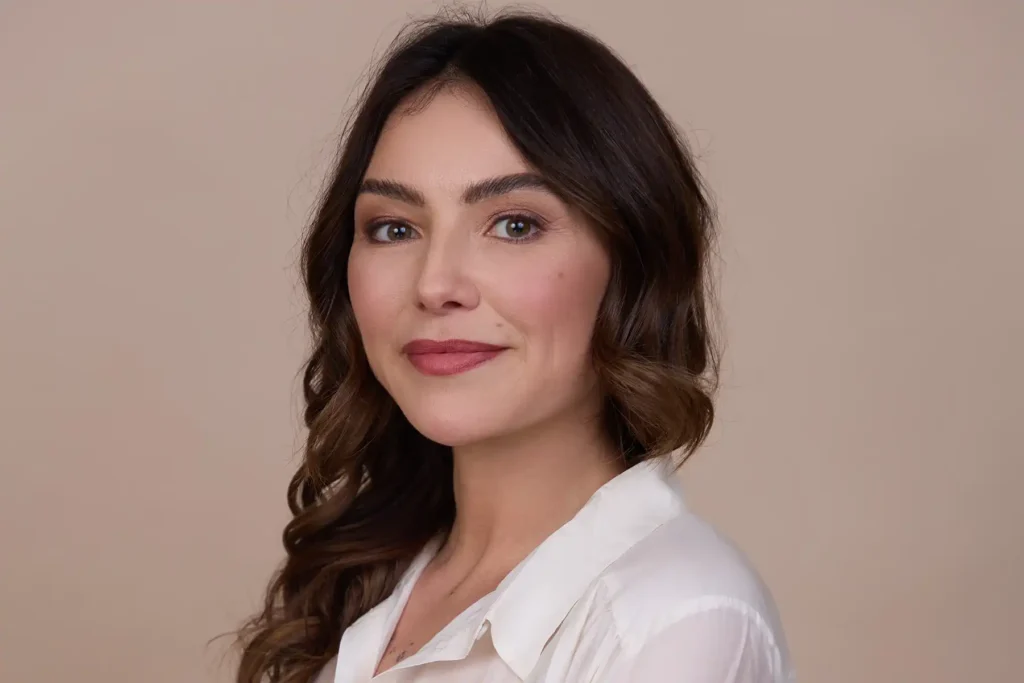 Nina, Heilpraktikerin bei Yaya prenatal, im Porträt vor beigem Studiohintergrund. Sie trägt eine weiße Bluse und lächelt ruhig in die Kamera. Ihre dunkelbraunen, welligen Haare fallen seitlich über die Schulter.