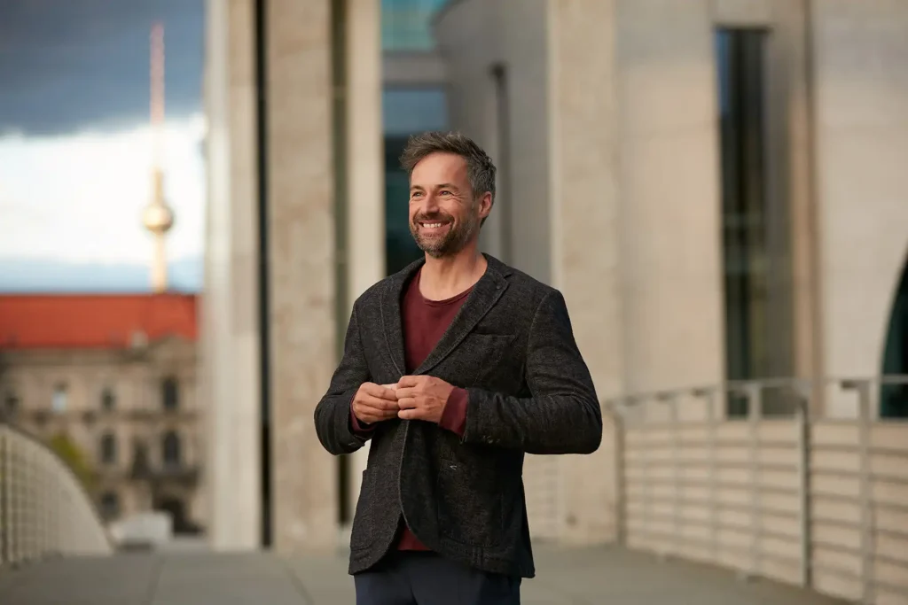 Outdoor Business Portraits Berlin eines lächelnden Mannes mittleren Alters mit kurzem, grau meliertem Haar und Bart. Er steht im Freien in Berlin und trägt einen dunklen Blazer über einem bordeauxroten T-Shirt. Sein Name ist Rayk. Er hält die Revers seines Blazers mit beiden Händen fest. Im verschwommenen Hintergrund sind die großen Steinsäulen eines modernen Gebäudes und der ikonische Berliner Fernsehturm in der Ferne zu sehen. Das Porträt wurde von Ed Mehravaran aufgenommen.