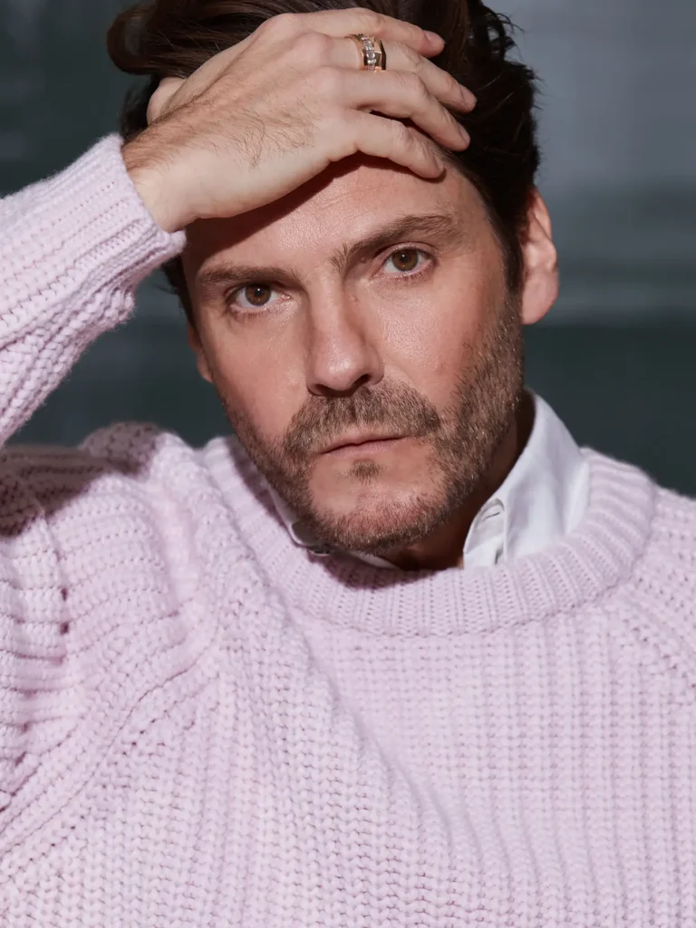 Schauspieler-Fotoshooting mit Daniel Brühl. Er trug einen hellrosa, grob gestrickten Zopfstrickpullover über einem weißen Hemd. Seine rechte Hand – mit einem goldenen Ring geschmückt – legte er an die Stirn, den Blick fest in die Kamera gerichtet. Der Hintergrund war in sanften Türkis- und Grautönen verschwommen. Das warme, gerichtete Licht warf sanfte Schatten auf sein Gesicht. Das Foto wurde von Ed Mehravaran in Berlin aufgenommen.