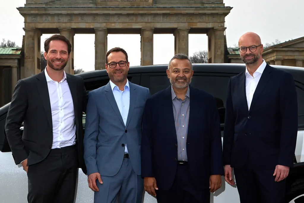 Businessporträt im Freien, aufgenommen vom Berliner Fotografen Ed Mehravaran. Vier Männer in Businesskleidung stehen lächelnd nebeneinander vor dem Brandenburger Tor in Berlin. Von links nach rechts: Frank Steuer im dunklen Anzug, Dr. Jens Wohltorf im hellblauen Anzug mit Brille, Pradeep Parameswaran in einer dunklen Jacke und ein weiterer Mann im dunklen Anzug mit Brille. Im Hintergrund ist ein dunkles Fahrzeug zu sehen.
