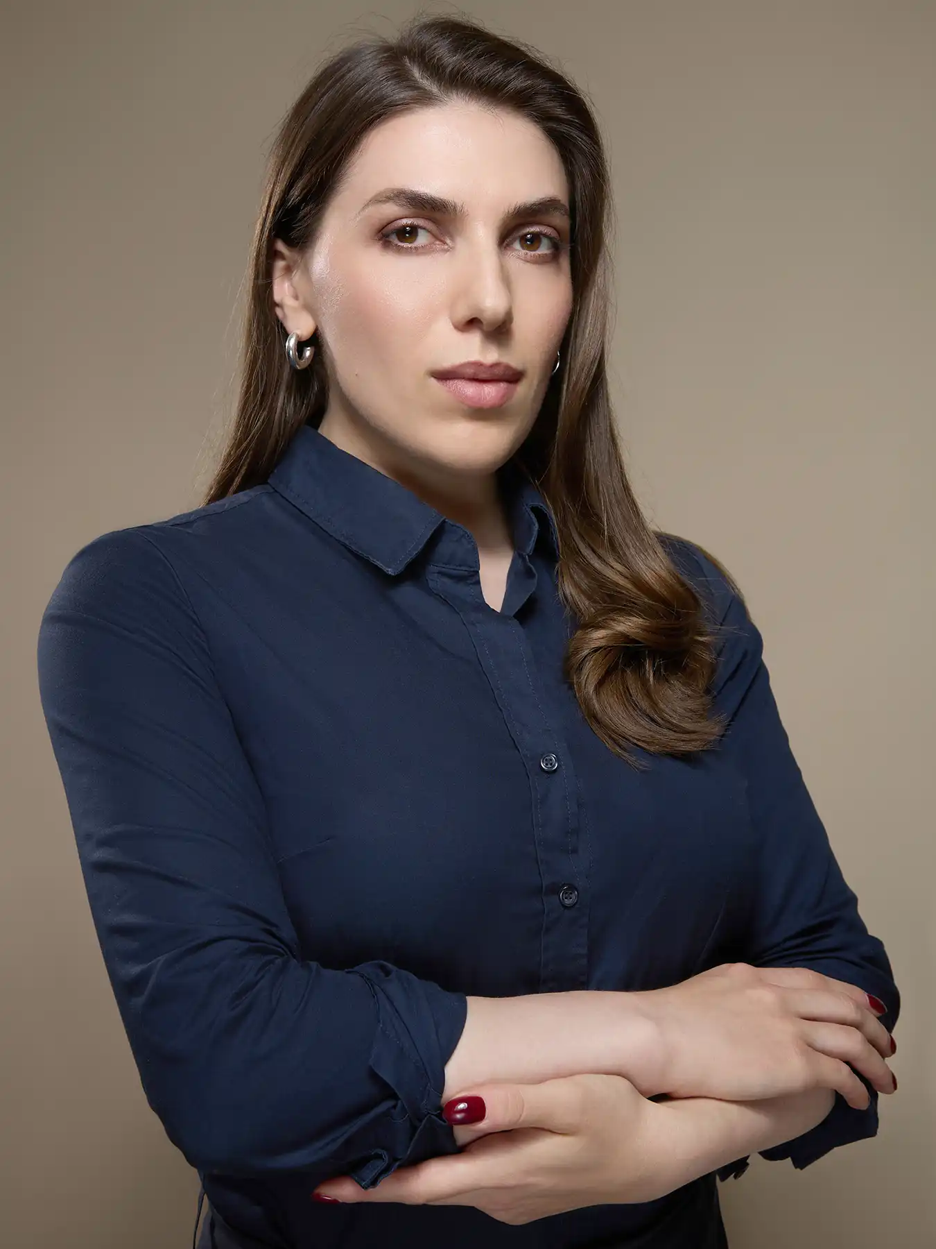 Lucy in a business portrait against a beige studio background. She is wearing a dark blue blouse and has her arms crossed. Her gaze is confident and directed straight into the camera. Silver hoop earrings and dark red painted nails complete the look. Her brown hair falls to the side over her shoulder.