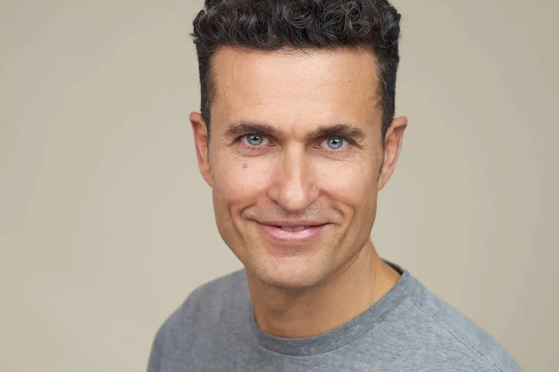 Claude responded: Gabriel Lazaro im Headshot-Portrait vor beigem Studiohintergrund.Gabriel Lazaro im Headshot-Portrait vor beigem Studiohintergrund. Er trägt ein graues T-Shirt und lächelt offen in die Kamera. Sein Blick ist durch markante blaue Augen und dunkle, kurze Haare geprägt.