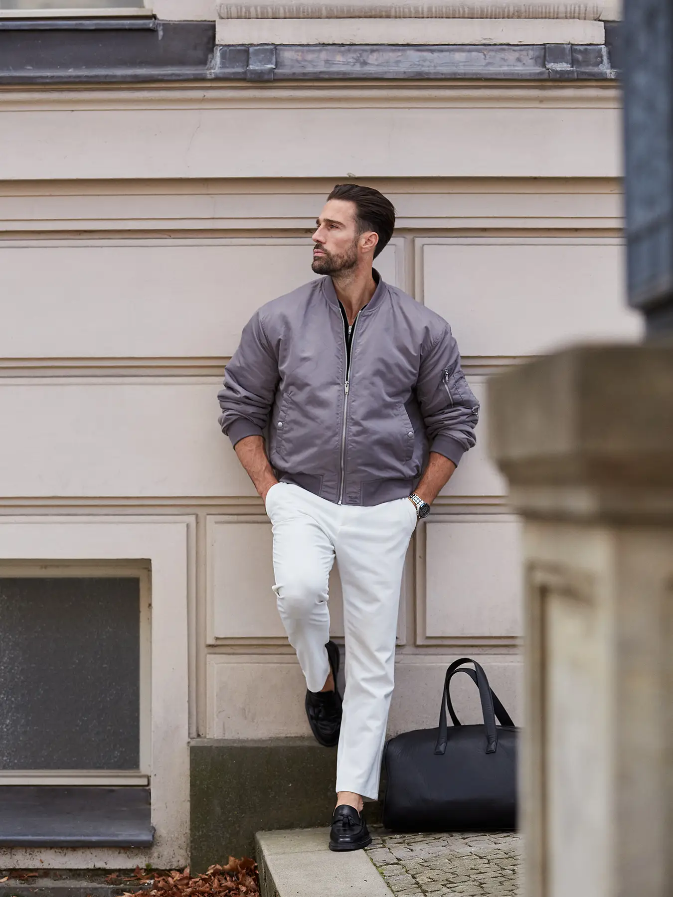 Goran lehnt lässig an einer hellen Altbaufassade in Berlin-Mitte. Er trägt eine graue Bomberjacke, weiße Chinos und schwarze Loafer. Neben ihm steht eine schwarze Leder-Tote-Bag. Sein Blick geht entspannt zur Seite.