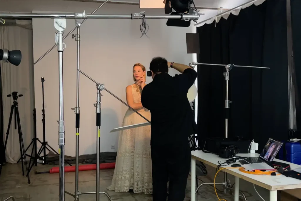 A behind-the-scenes look at a photo shoot in Ed Mehravaran's Berlin studio. A photographer dressed in black adjusts the camera position while a woman in a cream-colored lace dress poses against a bright background. Numerous tripods, flash units, and softboxes are set up all around her. On a table to the right, a laptop and a Canon camera are visible.