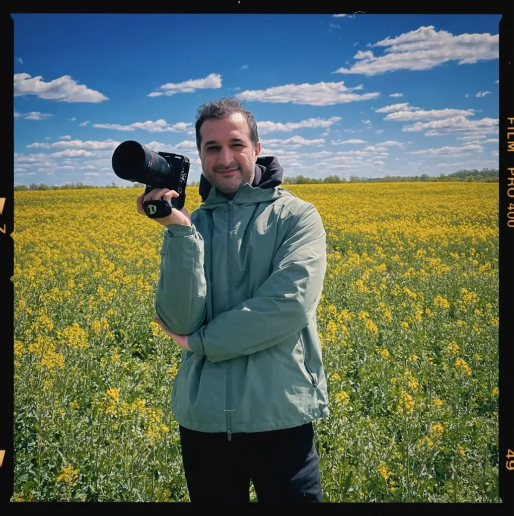 Ed Mehravaran mit grauem Bart und dunklen, leicht schütteren Haaren steht lächelnd in einem gelb blühenden Rapsfeld unter blauem Himmel mit lockeren Wolken. Er trägt eine mintgrüne Regenjacke und dunkle Hose und hält eine Kamera mit großem schwarzen Objektiv in der erhobenen rechten Hand. Das Bild hat einen Retro-Filmrand mit der Aufschrift „FILM PRO 400" am rechten Rand.