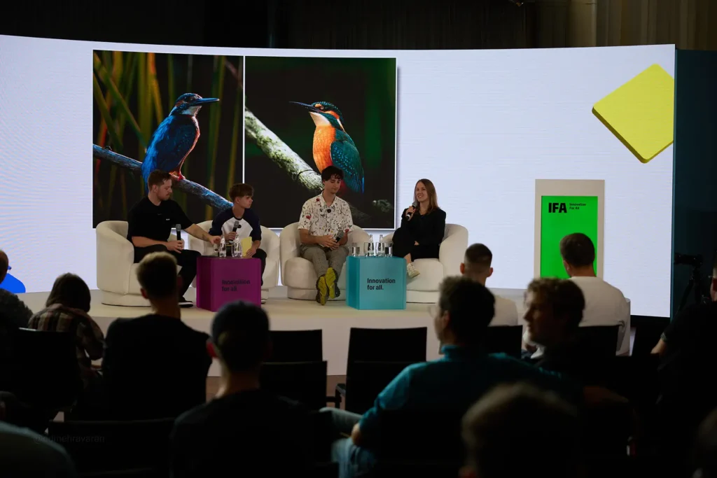 Panel discussion at IFA Berlin with three men and one woman seated in white armchairs on stage, holding microphones and speaking, in front of a large screen displaying two close-up photos of kingfisher birds and the "IFA Innovation for All" logo, with an audience seated in the foreground
