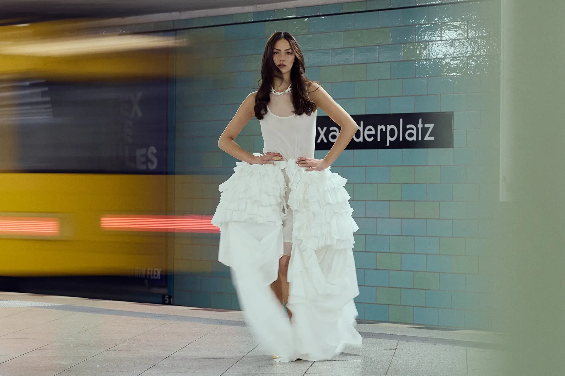 Ein Editorial-Modefoto, aufgenommen am Berliner U-Bahnhof Alexanderplatz von dem in Berlin ansässigen Profifotografen Ed Mehravaran: Eine Frau mit dunklem, welligem Haar posiert in einem komplett weißen Outfit – bestehend aus einem transparenten Oberteil und einem asymmetrischen Rock mit Rüschen – vor einer Kulisse aus türkisfarbenen Fliesen, während im Hintergrund ein gelber Zug vorbeifährt.