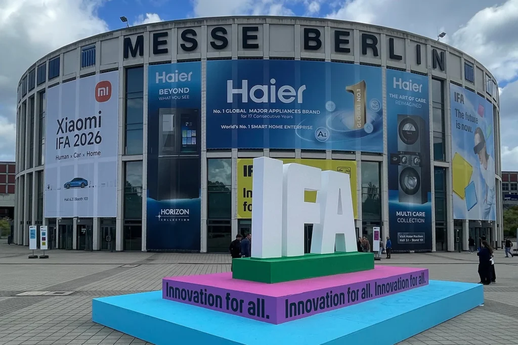Large white 'IFA' letters on a green, pink, and blue platform reading 'Innovation for all,' standing in front of the curved Messe Berlin building decorated with banners from Xiaomi and Haier for IFA 2026