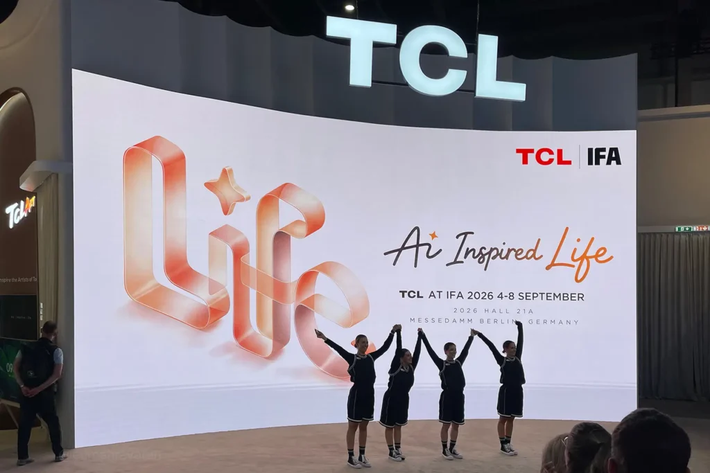 TCL booth at IFA Berlin 2026, with four performers in black outfits standing on stage holding hands raised, in front of a large curved screen displaying a stylized orange "Life" logo and the tagline "Ai Inspired Life," along with event details "TCL at IFA 2026, 4–8 September, Hall 21A, Messedamm Berlin, Germany.