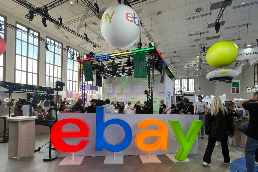 eBay booth at IFA Berlin 2026 with large colorful 3D "ebay" letters in the foreground, a giant eBay-branded balloon hanging overhead, and a live-streaming stage set up behind, with attendees and staff gathered around amid the busy exhibition hall.