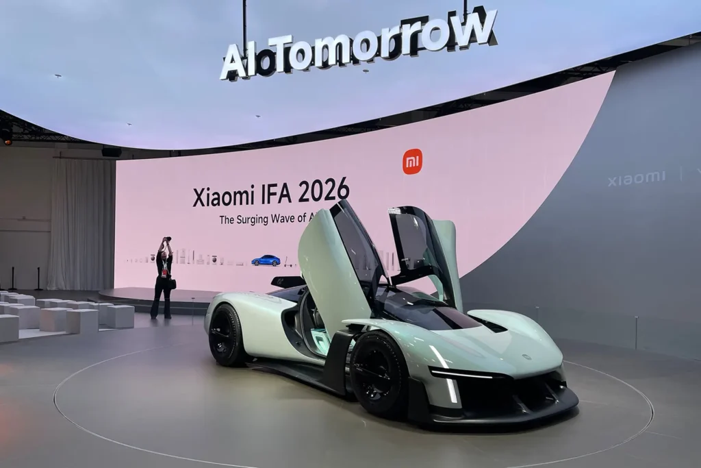 Xiaomi's IFA 2026 booth featuring a pale green concept sports car with upward-opening butterfly doors displayed on a rotating platform, in front of a large curved screen reading "Xiaomi IFA 2026, The Surging Wave of AI" with a "MI" logo, under an overhead "AIoTomorrow" sign, with an attendee photographing the display in the background
