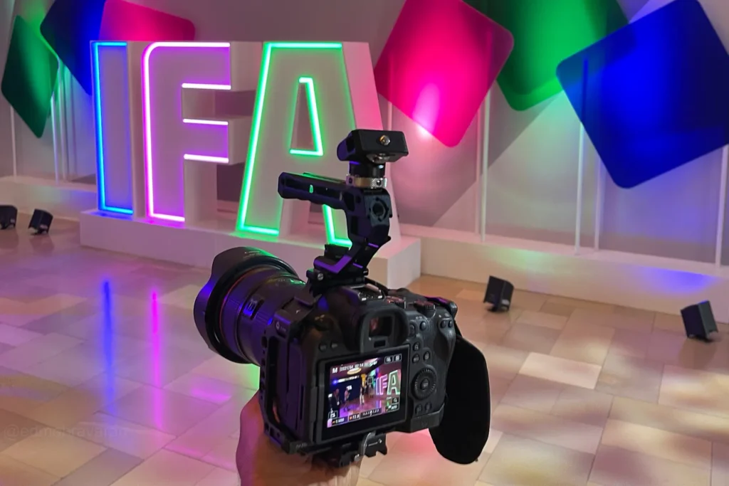 A mirrorless camera with a rig and handle in the foreground, its screen showing a live shot of people on stage, with a glowing neon 'IFA' sign and colorful geometric panels lit in pink, green, and blue in the background