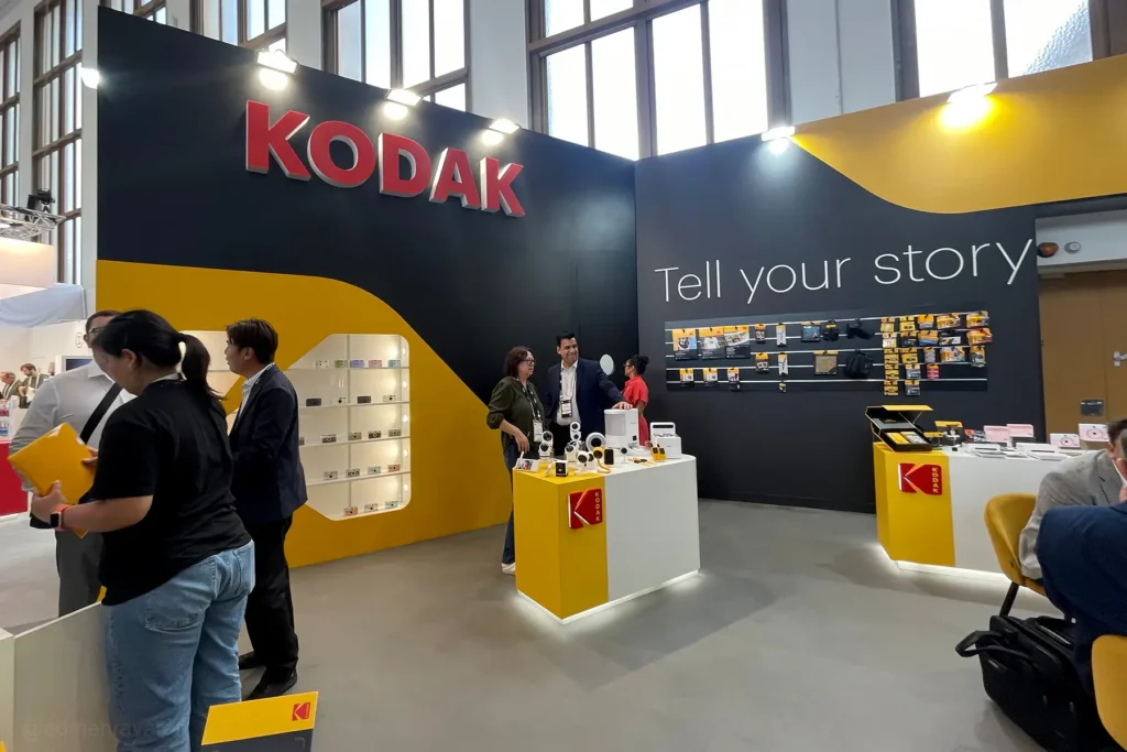 Kodak trade fair booth at IFA Berlin with a large red "KODAK" logo and "Tell your story" tagline on dark walls, featuring yellow display counters with cameras and accessories, a lit shelf of point-and-shoot cameras, and several attendees browsing and chatting near the exhibits