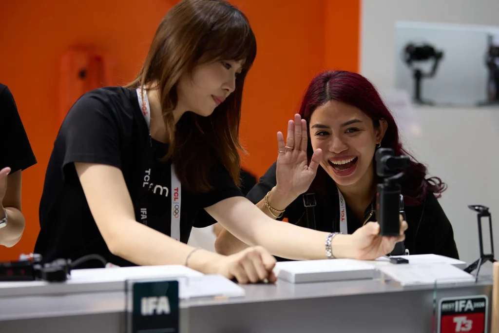 Two women at a TCL exhibition booth, one demonstrating a handheld gimbal camera while the other waves and laughs, with cameras displayed on stands in the background and an IFA award sign on the counter